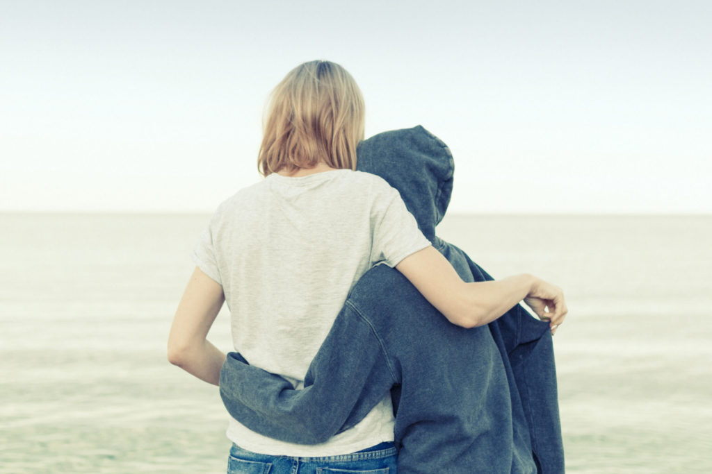 Adult and teenager embracing by sea with backs to us